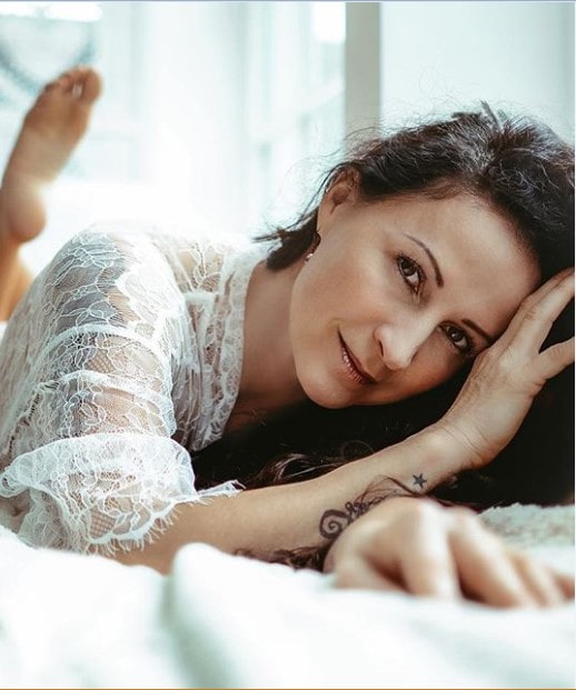 Fotograf, zgodna kovrdžava žena pozira na krevetu u providnoj beloj majici