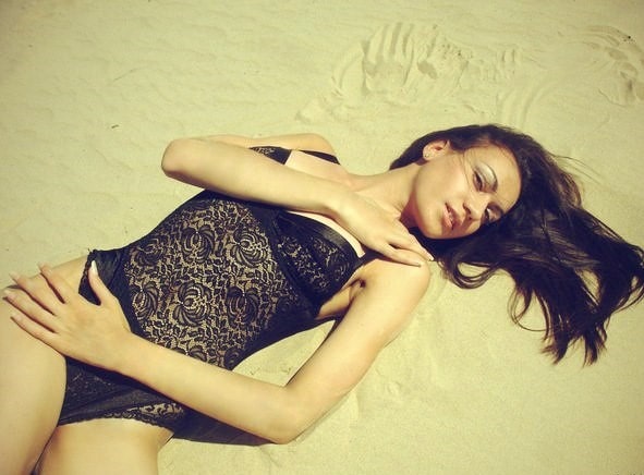 Nasmejana lepa crnka pozira na pesku na plaži u čipkanom crnom kupaćem kostimu