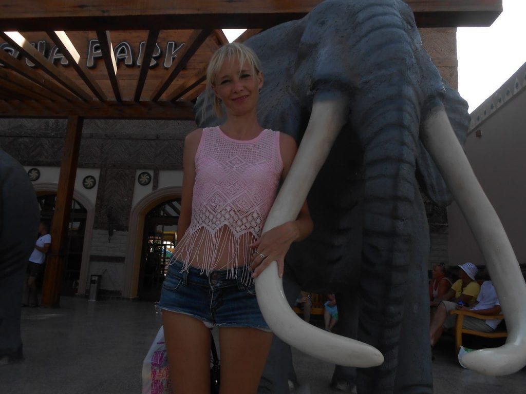 Situirana lepa plavuša pozira u parku držeči se za figuru slona