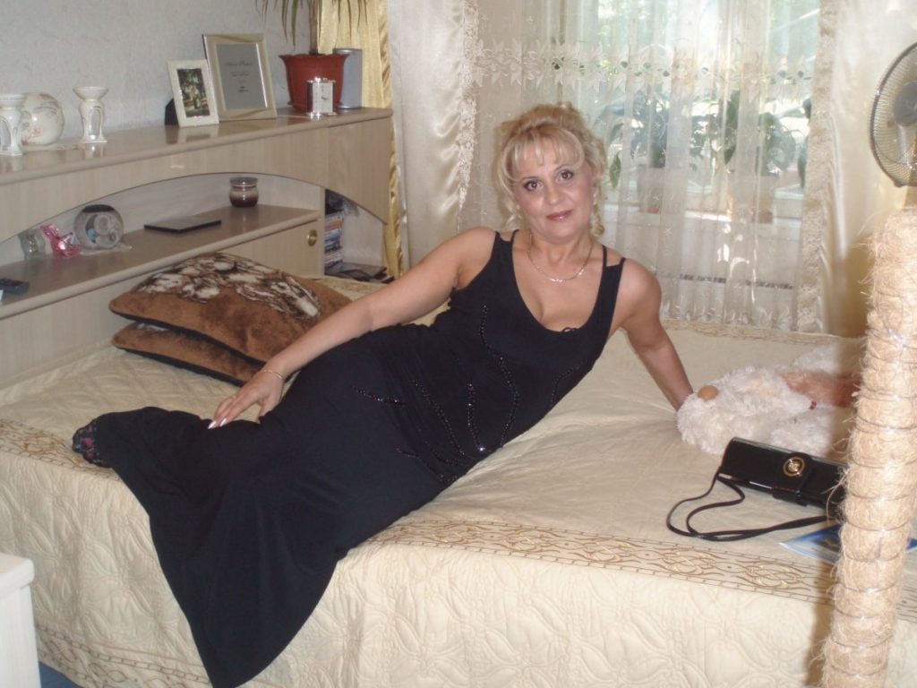 Iskrena, lepa plavuša u godinama pozira provokativno na krevetu u dugoj crnoj haljini